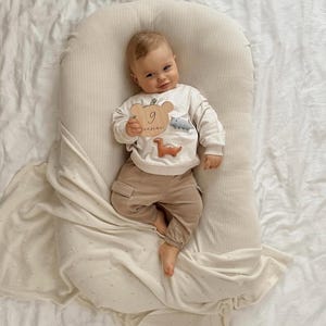 May include: A baby resting in a cream-colored ribbed lounger, wearing a long-sleeved cream sweatshirt with dinosaur appliqu&eacute;s and tan cargo pants. The baby holds a wooden bear-shaped milestone plaque with the number 9 and cursive text.
