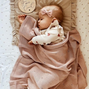May include: A dusty rose knitted baby blanket with a delicate pointelle heart pattern, a beige ruffled pillow with a wooden disc reading You Are So Loved, and a baby wearing a cream long-sleeve shirt with red hearts and a small pink bow headband.