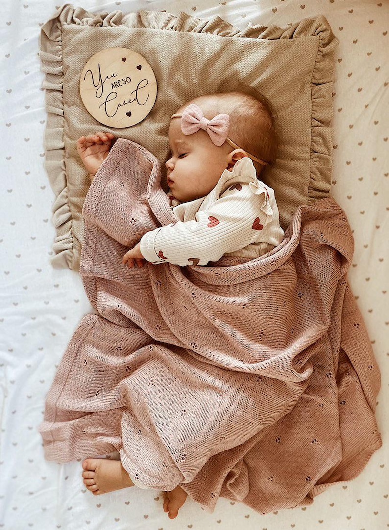 May include: A dusty rose knitted baby blanket with a delicate pointelle heart pattern, a beige ruffled pillow with a wooden disc reading You Are So Loved, and a baby wearing a cream long-sleeve shirt with red hearts and a small pink bow headband.