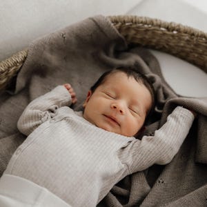 May include: A newborn infant with dark hair sleeping peacefully while wearing a light beige ribbed long-sleeve bodysuit. The infant is resting inside a woven wicker basket lined with a soft, dark taupe knit blanket featuring a subtle pointelle pattern.