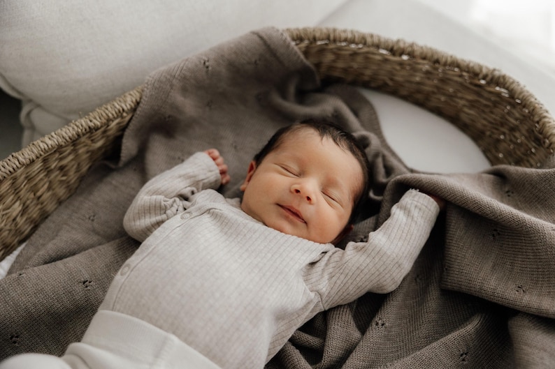 May include: A newborn infant with dark hair sleeping peacefully while wearing a light beige ribbed long-sleeve bodysuit. The infant is resting inside a woven wicker basket lined with a soft, dark taupe knit blanket featuring a subtle pointelle pattern.