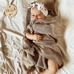 May include: A baby wrapped in a light brown knit blanket with a delicate eyelet pattern. The baby wears a white headband featuring a small cherry print. A circular wooden disc nearby is engraved with the text Oh Girl and small heart icons.