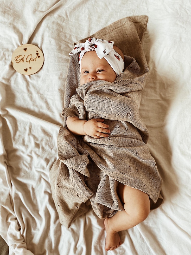May include: A baby wrapped in a light brown knit blanket with a delicate eyelet pattern. The baby wears a white headband featuring a small cherry print. A circular wooden disc nearby is engraved with the text Oh Girl and small heart icons.