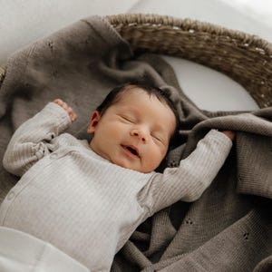May include: A newborn infant with dark hair is sleeping peacefully inside a woven wicker basket. The infant is wearing a long-sleeved, ribbed, light beige bodysuit and is partially covered by a soft, taupe-colored knit blanket with a delicate eyelet pattern.
