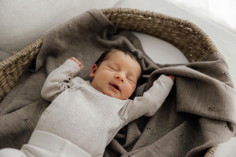 May include: A newborn infant with dark hair is sleeping peacefully inside a woven wicker basket. The infant is wearing a long-sleeved, ribbed, light beige bodysuit and is partially covered by a soft, taupe-colored knit blanket with a delicate eyelet pattern.