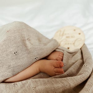 May include: A beige knitted baby blanket with a delicate pointelle pattern featuring small eyelet details. The blanket is draped over a pair of small feet. A round wooden disc is visible with the engraved text "You are so loved" in script.