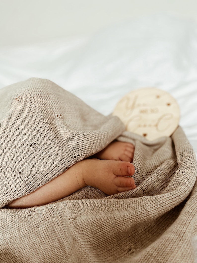 May include: A beige knitted baby blanket with a delicate pointelle pattern featuring small eyelet details. The blanket is draped over a pair of small feet. A round wooden disc is visible with the engraved text "You are so loved" in script.