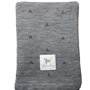 May include: A rectangular grey knitted blanket featuring a repeating pattern of small, three-hole eyelet details. A white rectangular fabric label is sewn onto the center, displaying a gold butterfly graphic and the text Blatterschwing.