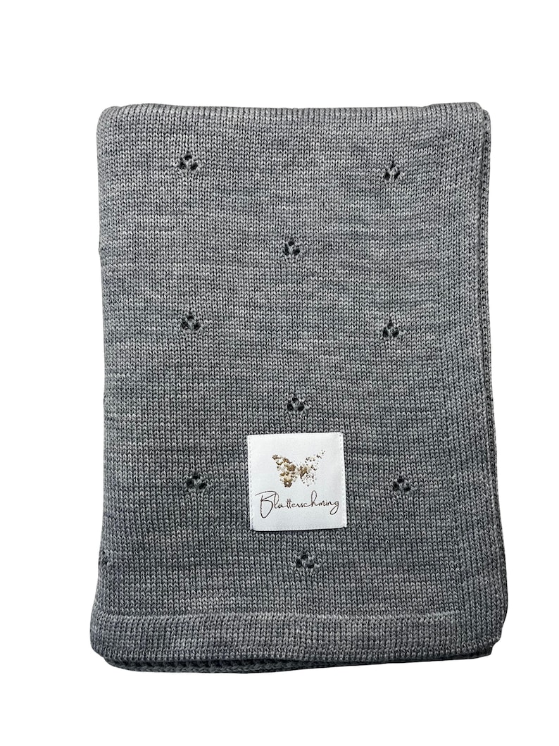 May include: A rectangular grey knitted blanket featuring a repeating pattern of small, three-hole eyelet details. A white rectangular fabric label is sewn onto the center, displaying a gold butterfly graphic and the text Blatterschwing.