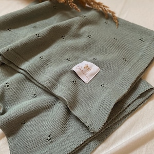 May include: A sage green knitted blanket featuring a delicate pointelle pattern of small, evenly spaced eyelet holes. A white fabric label is sewn onto the surface, displaying a butterfly graphic and the text Bl&auml;tterschwing in cursive script.