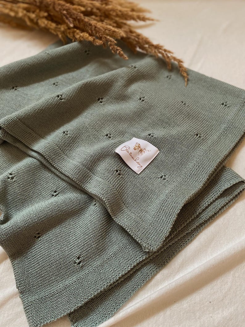 May include: A sage green knitted blanket featuring a delicate pointelle pattern of small, evenly spaced eyelet holes. A white fabric label is sewn onto the surface, displaying a butterfly graphic and the text Bl&auml;tterschwing in cursive script.
