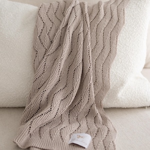 Peut inclure: Une couverture beige tricotée avec un motif ondulé est drapée sur un oreiller blanc. La couverture a une étiquette attachée. La couverture est faite d'un matériau doux, probablement pour la chaleur et le confort. Dimensions en centimètres.