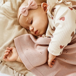 May include: A sleeping infant wearing a cream-colored long-sleeve shirt with a red heart pattern, a dusty pink knit blanket with a subtle eyelet design, and a dusty pink fabric bow headband on a thin elastic band.