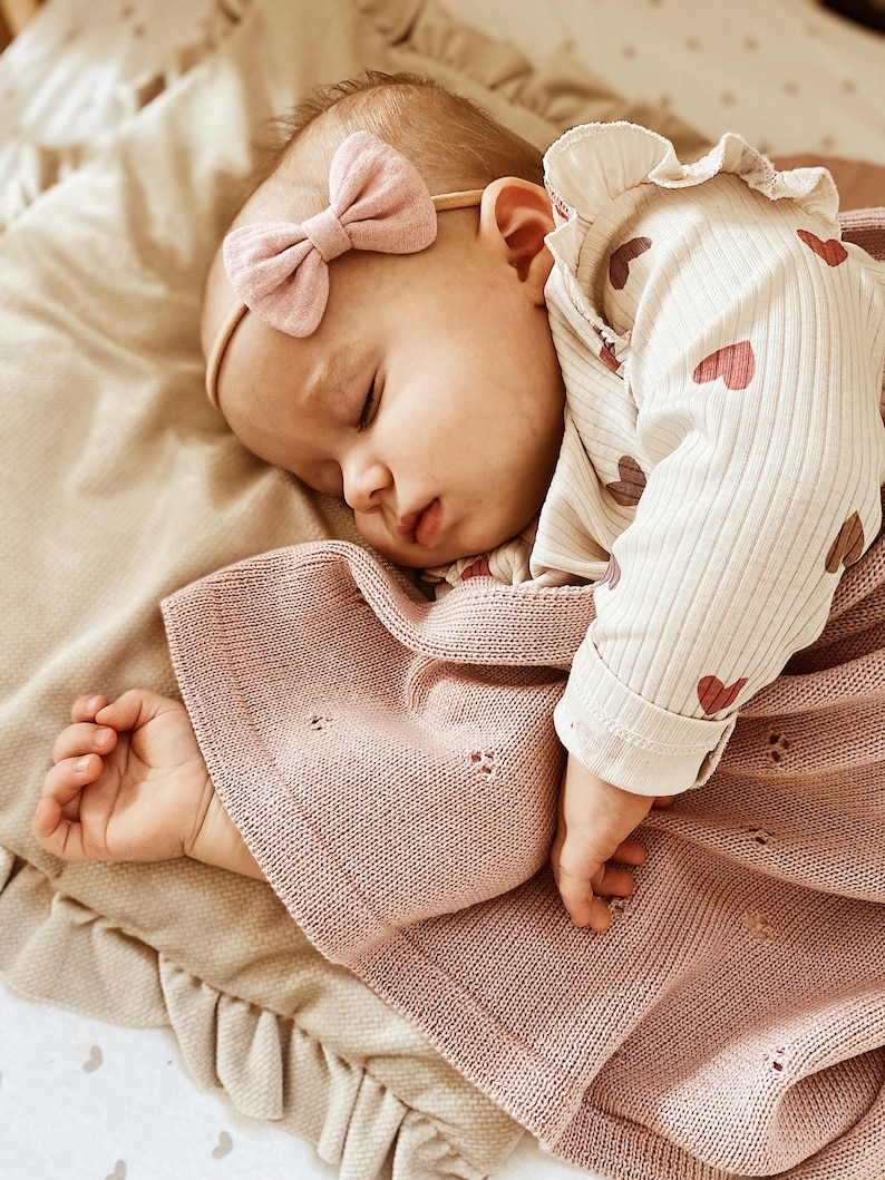 May include: A sleeping infant wearing a cream-colored long-sleeve shirt with a red heart pattern, a dusty pink knit blanket with a subtle eyelet design, and a dusty pink fabric bow headband on a thin elastic band.