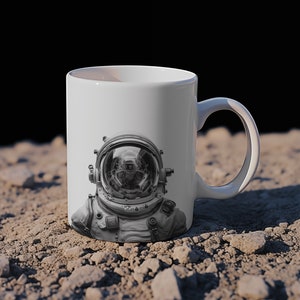 May include: White ceramic mug with a black and white image of an astronaut wearing a helmet. The mug is sitting on a gray, rocky surface.