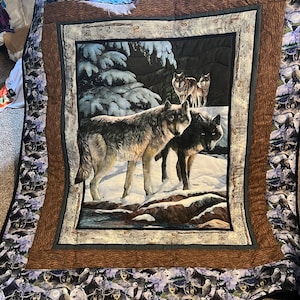 May include: A large, rectangular quilt featuring a central image of wolves in a snowy landscape. The quilt has a brown border and a patterned edge with images of wolves and birds. The central image depicts several wolves in a winter scene.