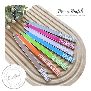 Könnte beinhalten: Bunte Gartenstecker mit Pflanzennamen in weißer Schrift. Die Stecker sind auf einem Holzbrett angeordnet. Der Text "Mix & Match" und "SO INDIVIDUELL WIE DEIN GARTEN" sind ebenfalls sichtbar.