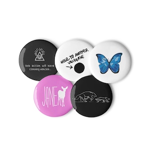 May include: Set of five black and white button badges with various designs. One badge features a white deer with the word "JANE" in white text. Another badge features a black and white illustration of a wolf chasing a smaller wolf. A third badge features a black and white illustration of an eye with a triangle above it and the text "this action will have consequences..." A fourth badge features a black circle with a white arrow pointing to it and the text "HOLE TO ANOTHER UNIVERSE". The fifth badge features a blue butterfly on a white background.
