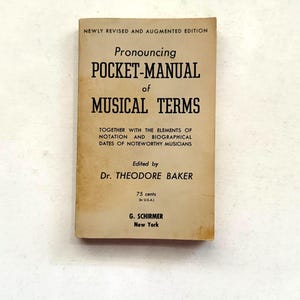 May include: A vintage pocket-sized book titled "Pronouncing Pocket-Manual of Musical Terms." The cover is tan with black text, including the author's name, Dr. Theodore Baker, and the publisher, G. Schirmer, New York. The book is a guide to musical terms.