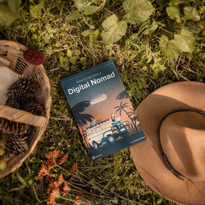 May include: A book titled "How to Become a Digital Nomad" by Amanda Gauld, lying on a bed of green grass with a brown hat and a wicker basket with pine cones.