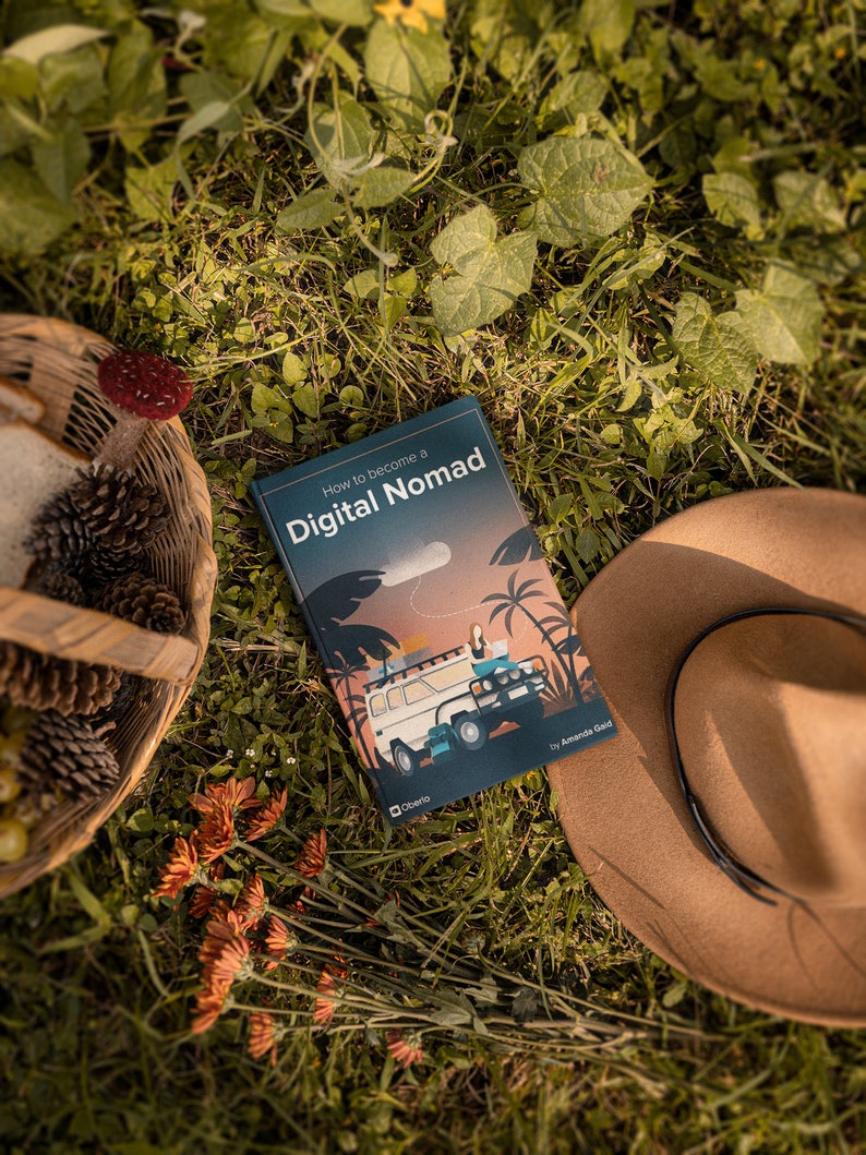 May include: A book titled "How to Become a Digital Nomad" by Amanda Gauld, lying on a bed of green grass with a brown hat and a wicker basket with pine cones.