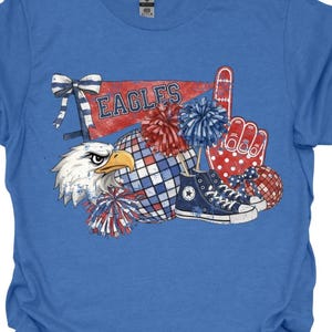 May include: Blue t-shirt featuring a graphic design with the word "EAGLES" on a pennant, an eagle head, pom-poms, a foam hand, and a pair of blue sneakers. The design incorporates red, white, and blue colors.