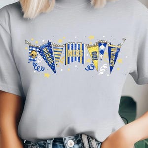 May include: Light gray t-shirt featuring a banner design with pennants in blue, yellow, and white. The pennants display text and a wildcat logo. The design includes stars and decorative swirls, creating a festive look.