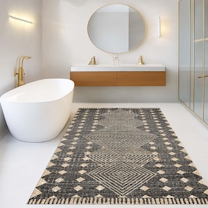 Puede incluir: Una alfombra rectangular de baño con un patrón geométrico de diamantes en negro, gris y beige. La alfombra está colocada sobre un suelo blanco en un baño moderno, con una bañera blanca y accesorios dorados.