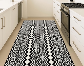 May include: A black and white geometric patterned rug in a kitchen setting. The rug features a repeating design of zigzags, triangles, and diamond shapes. The rug is rectangular and appears to be made of a durable material, suitable for high-traffic areas.