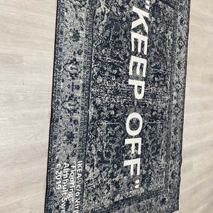 May include: Black and white rug with a vintage pattern and the text "KEEP OFF" in white. The rug is labeled "IKEA&reg; c/o Virgil Abloh" and "Flooring" in white text. The rug is made in Almhult, Sweden.