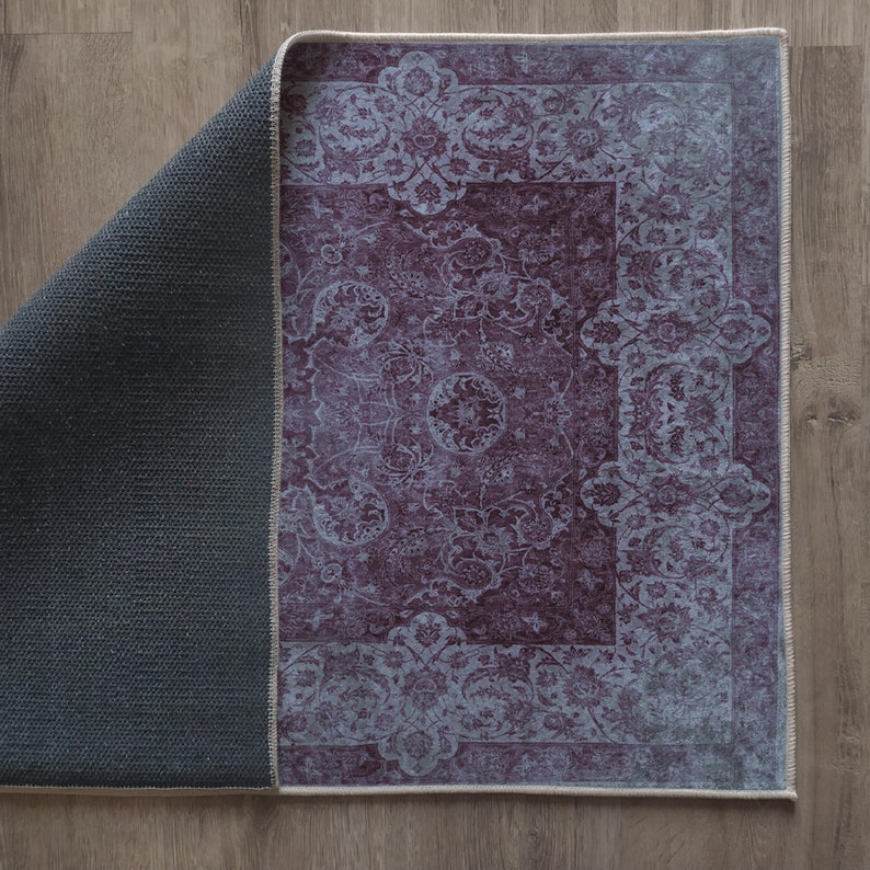 May include: A purple and white patterned rug with a fringed edge. The rug is folded in half, showing the dark gray backing.