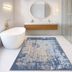 May include: A rectangular rug with a distressed pattern in blue, beige, and grey. The rug is placed on a white floor in a bathroom, with a white bathtub, gold fixtures, and a wooden vanity in the background. The rug's dimensions are approximately 170cm x 240cm.