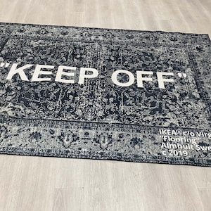 May include: A black and white rug with a vintage pattern. The rug has the text "KEEP OFF" in white letters. The rug is labeled "IKEA c/o Virgil Abloh "Flooring" Almhult Sweden C 2019".