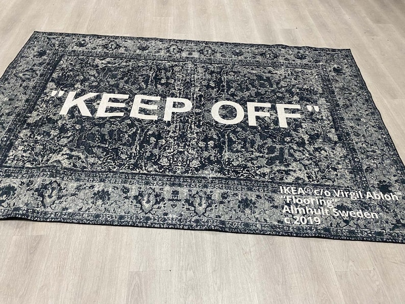 May include: A black and white rug with a vintage pattern. The rug has the text "KEEP OFF" in white letters. The rug is labeled "IKEA c/o Virgil Abloh "Flooring" Almhult Sweden C 2019".