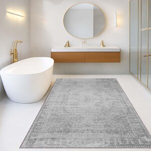 May include: A gray rectangular rug with an ornate, faded pattern is centered in a modern bathroom. The rug is placed in front of a white bathtub and a wooden vanity with a gold-framed mirror. The bathroom has white walls and gold fixtures.