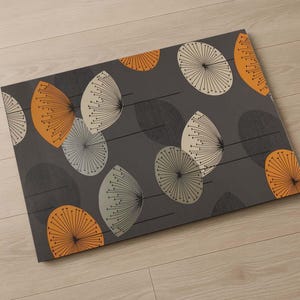 Op de afbeelding: Een rechthoekig grijs tapijt met een herhaald patroon van gestileerde paardenbloemzaden in oranje, crème en grijs. Het tapijt heeft een modern, geometrisch ontwerp, geschikt voor een woonkamer of entree.