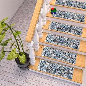 Puede incluir: Un conjunto de cinco peldaños antideslizantes para escaleras con un diseño de piedras de guijarro. Los peldaños son de color beige con piedras grises y azules. Los peldaños están en una escalera de madera con una barandilla blanca.
