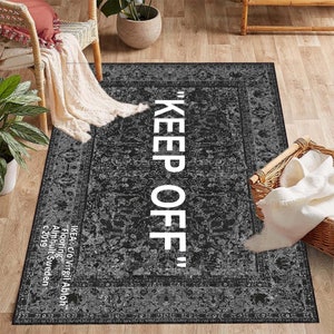 May include: Black and white area rug with a vintage design and the text "KEEP OFF" in white. The rug has a border with a floral pattern. The text "IKEA@c/o Virgil Abloh" is printed in white at the bottom of the rug.