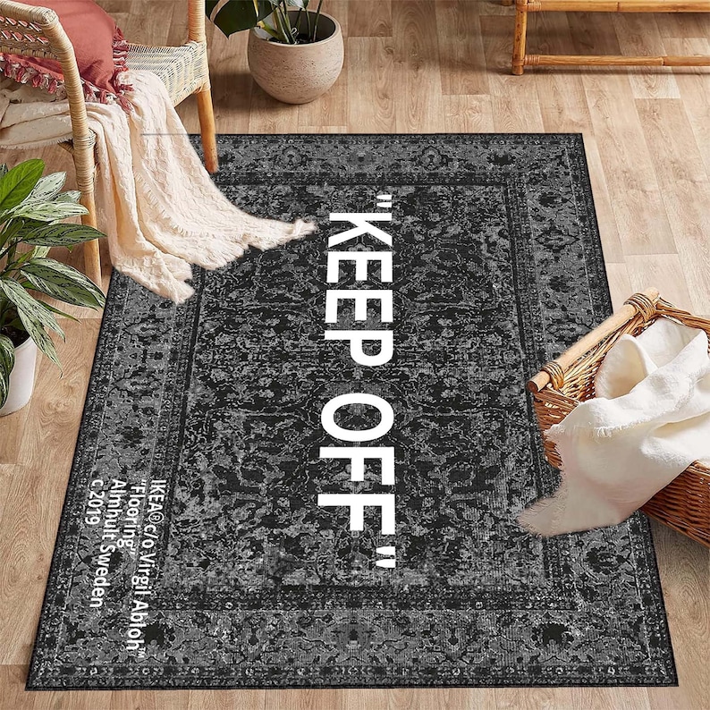 May include: Black and white area rug with a vintage design and the text "KEEP OFF" in white. The rug has a border with a floral pattern. The text "IKEA@c/o Virgil Abloh" is printed in white at the bottom of the rug.