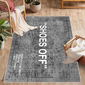 Könnte beinhalten: Grauer rechteckiger Teppich mit floralem Muster und dem Schriftzug "SHOES OFF" in großen weißen Buchstaben. Der Teppich hat einen Used-Look und liegt auf einem Holzboden. Teil der IKEA c/o Virgil Abloh "Flooring" Kollektion.