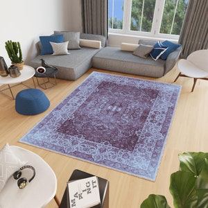 May include: A vintage-style rug with a light blue and purple color scheme. The rug has a floral pattern and a border with a repeating design. The rug is laid out on a wooden floor in a living room setting.