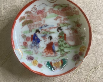 May include: A small, round, white ceramic dish with a red rim. The dish features a hand-painted scene of figures in traditional Japanese attire, a landscape, and a butterfly. The dish is decorated with gold accents.