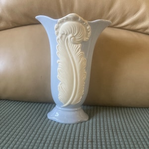 May include: A light blue ceramic vase with a white, raised feather design. The vase has a flared top and a pedestal base. The feather design is centered on the front of the vase. The vase is approximately 8 inches tall.