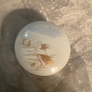 May include: A white ceramic trinket box with a seashell and starfish design. The box has a blue and white sky design with gold accents.