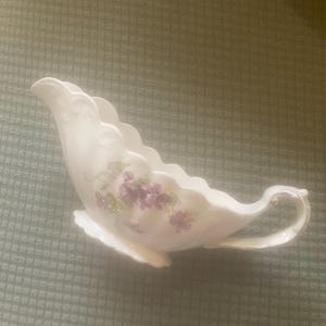 May include: A white porcelain gravy boat with a scalloped rim and a curved handle. The boat features a floral design with purple flowers and green leaves. The gravy boat is on a green surface.