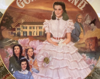 Peut inclure: Une assiette en porcelaine avec un bord doré présente une peinture de Scarlett O'Hara du film Autant en emporte le vent. L'assiette porte le titre "Autant en emporte le vent" en lettres dorées. L'arrière-plan de l'assiette est une scène de coucher de soleil avec une grande maison et un champ de fleurs.
