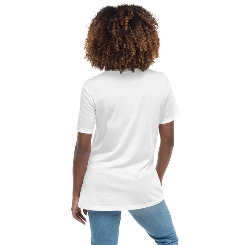 May include: A white t-shirt with a crew neck and short sleeves. The shirt is paired with blue jeans. The person in the image has curly brown hair.