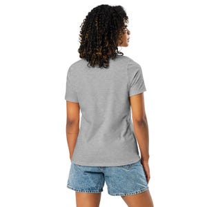May include: A grey t-shirt with short sleeves and denim shorts. The model is seen from the back, displaying the t-shirt and shorts. The outfit is casual and suitable for everyday wear. The t-shirt is a light shade of grey.