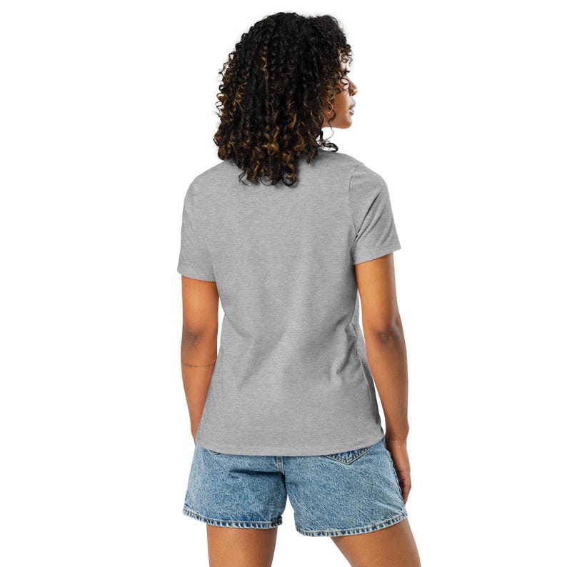 May include: A grey t-shirt with short sleeves and denim shorts. The model is seen from the back, displaying the t-shirt and shorts. The outfit is casual and suitable for everyday wear. The t-shirt is a light shade of grey.