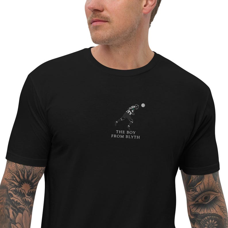 May include: Black t-shirt featuring a minimalist illustration of a football player throwing a ball. The text "THE BOY FROM BLYTH" is printed below the graphic in a simple font. The shirt is made of a soft material.